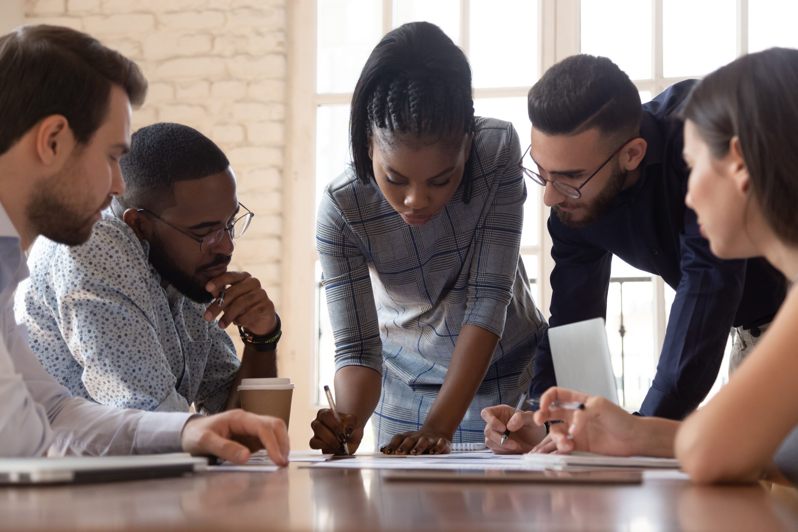 Focused arican american millennial female team leader leaned over table, writing down project ideas, editing documentation at brainstorming business meeting with diverse partners or clients at office.