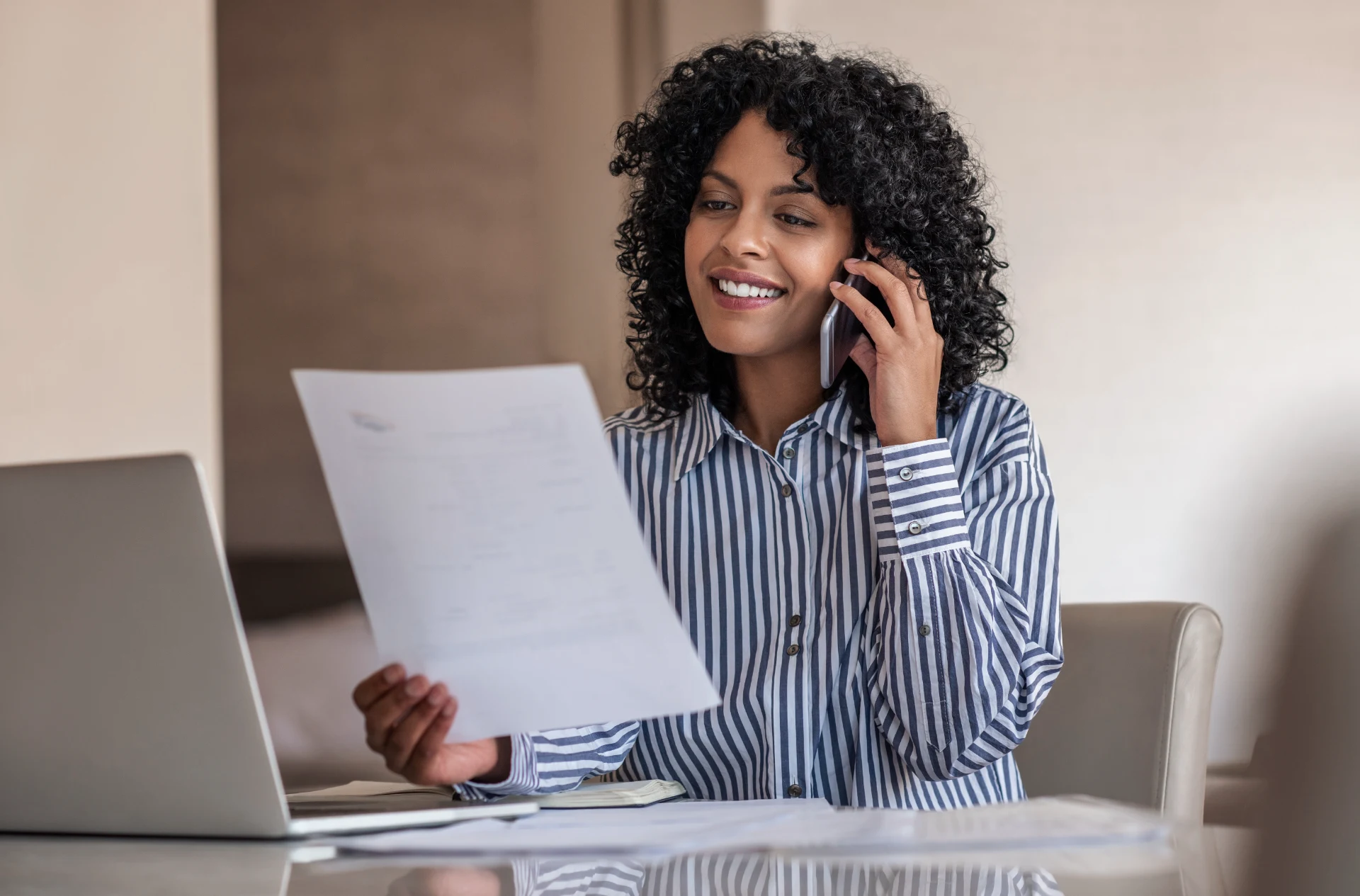 Smiling young female entrepreneur reading a document and talking with a client on her cellphone while working on a laptop at home