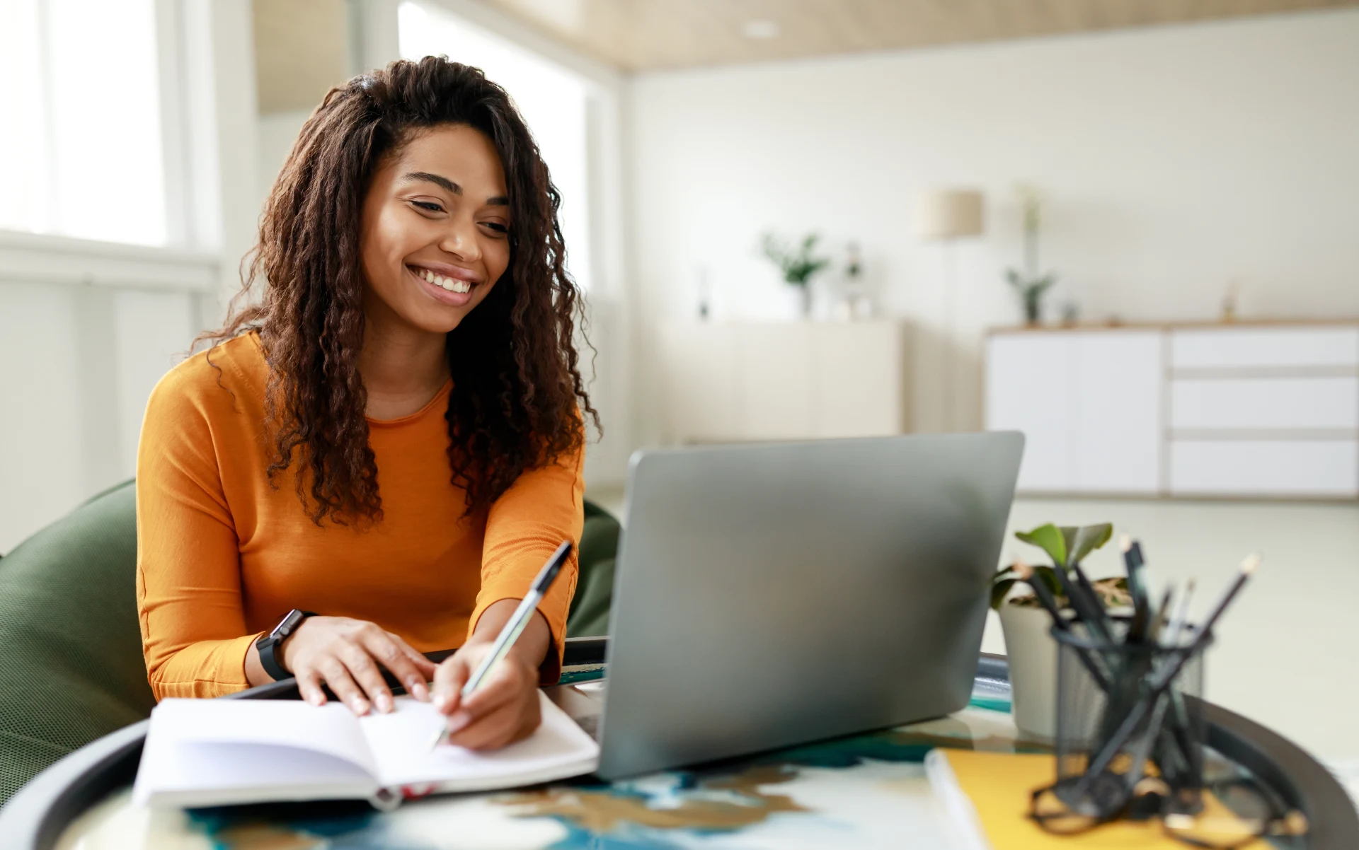 Smiling young African American woman sitting at desk working on laptop taking notes in notebook, happy millennial female studying online, watching webinar using computer and writing check list